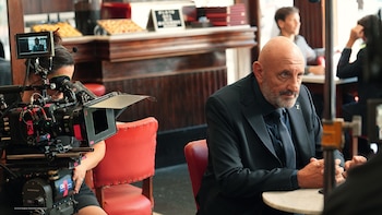 Un primer plano de un cameraman operando una Steadicam Arri en una cafetería, filmando a un hombre calvo con barba y traje sentado en una mesa
