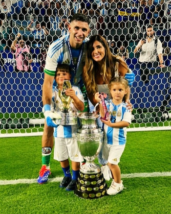 El futbolista Emiliano Martínez y su pareja Mandinha Martínez posan sonrientes con sus dos hijos y el trofeo de la Copa América en un campo de fútbol