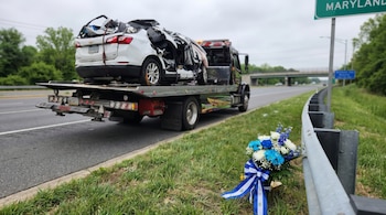 Un vehículo blanco severamente dañado es remolcado en una autopista en Maryland. Un ramo de flores azules y blancas con un lazo salvadoreño yace en la hierba.