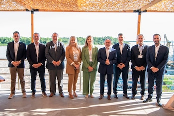 Nueve personas, siete hombres y dos mujeres, posan sonrientes al aire libre. Visten traje de negocios, con un río, vegetación y un muelle al fondo bajo un toldo