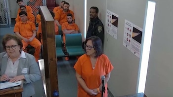 Una mujer con gafas y uniforme naranja frente a un micrófono, junto a una abogada, un guardia y varios detenidos sentados al fondo
