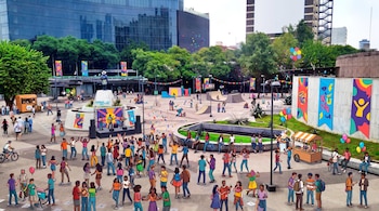 Una plaza urbana con un gran grupo de jóvenes, un concierto en vivo, rampas de patineta, puestos de venta, globos y banderines decorativos, rodeada de edificios.