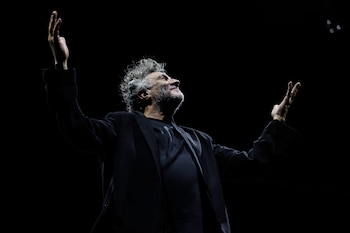 Fito Páez de perfil, con cabello rizado y gris, barba, brazos levantados y la cabeza inclinada hacia atrás, iluminado por un foco contra un fondo negro