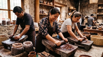 Tres personas muelen granos de cacao con rodillos sobre metates de piedra en una mesa con recipientes de barro y bolsas de semillas.