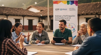 Grupo de hombres y mujeres de diversas edades sentados en una mesa de madera al aire libre, conversando y sonriendo. Detrás, un cartel de IGM PNUD.