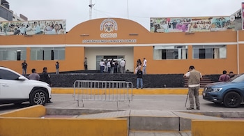 Edificio municipal naranja con logo de Comas, personas en la entrada, automóvil blanco, automóvil azul, barreras de seguridad, pavimento.