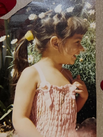Fotografía de una niña sonriendo de perfil, con cabello castaño recogido en coleta con banda amarilla, y top rosa smock, frente a un jardín de margaritas