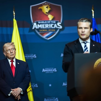 Donald Trump, con traje azul y corbata roja, de pie frente a una bandera amarilla y un logo de 'Shield of the Americas'. Un orador en un podio desenfocado
