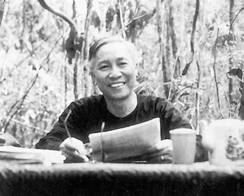 Fotografía en blanco y negro de un hombre sonriente sentado ante una mesa de madera con papeles, gafas, una taza y una tetera en el bosque