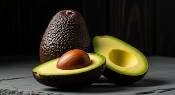 Aguacate Hass entero y dos mitades de aguacate Hass, una con hueso marrón y otra sin hueso, sobre una superficie de piedra gris oscura con fondo negro.
