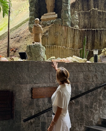 Una mujer de espaldas, con vestimenta blanca, extiende la mano hacia estatuas de piedra en una gruta con placas de texto, cactus y una palmera