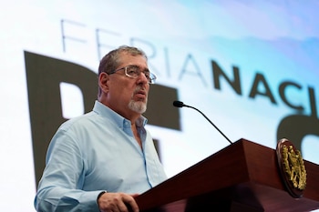 Bernardo Arévalo habla frente a un micrófono en un podio de madera con el escudo de Guatemala y una pantalla al fondo