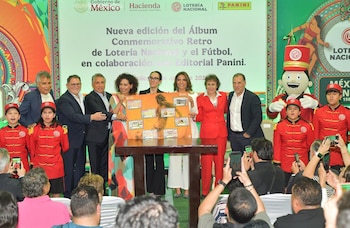 Un grupo de adultos y niños en uniformes rojos posan con un cartel del "Álbum Conmemorativo Retro de Lotería Nacional y el Fútbol" durante su lanzamiento