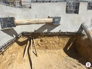 Fosa de excavación con paredes de concreto y armazones de metal, y un gran tronco de madera horizontal que atraviesa una pared. Se ven soportes de madera en la tierra