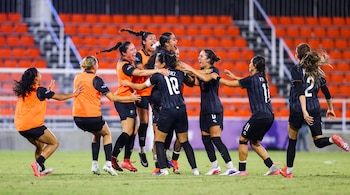 Varias jugadoras de fútbol femenino celebran en un campo. Llevan uniformes naranja y negro. Hay gradas vacías en el fondo.