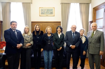 Siete personas, tres hombres y cuatro mujeres, posan de pie en una sala formal con paneles de madera, ventanas grandes y cortinas claras