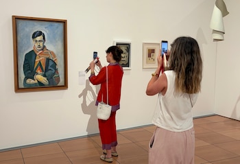 Dos mujeres en una galería de arte. Una pintura de un hombre con bufanda. Otros dos cuadros enmarcados y una escultura blanca. Paredes claras y suelo de baldosas marrones