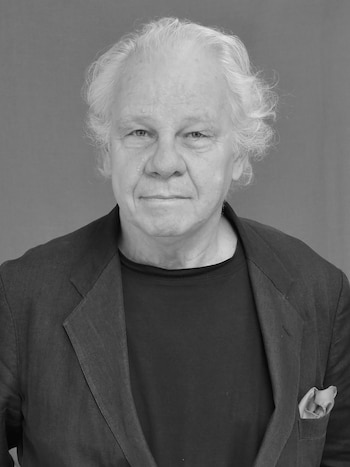 Retrato en blanco y negro de un hombre con pelo blanco, camisa oscura, chaqueta oscura y pañuelo de bolsillo. Fondo liso gris