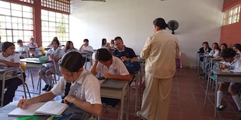 Estudiantes en uniforme sentados en pupitres escribiendo en sus cuadernos mientras una profesora y un hombre observan la clase