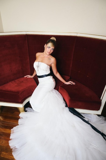 Marta Fort usa vestido branco com decote tomara que caia, laço preto e saia de tule, sentada em um sofá de veludo vermelho. O cabelo dela está amarrado