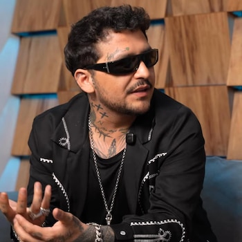 Primer plano de Peso Pluma con gafas de sol oscuras, chaqueta negra con bordado blanco, y tatuajes en el cuello y la cara, hablando y gesticulando