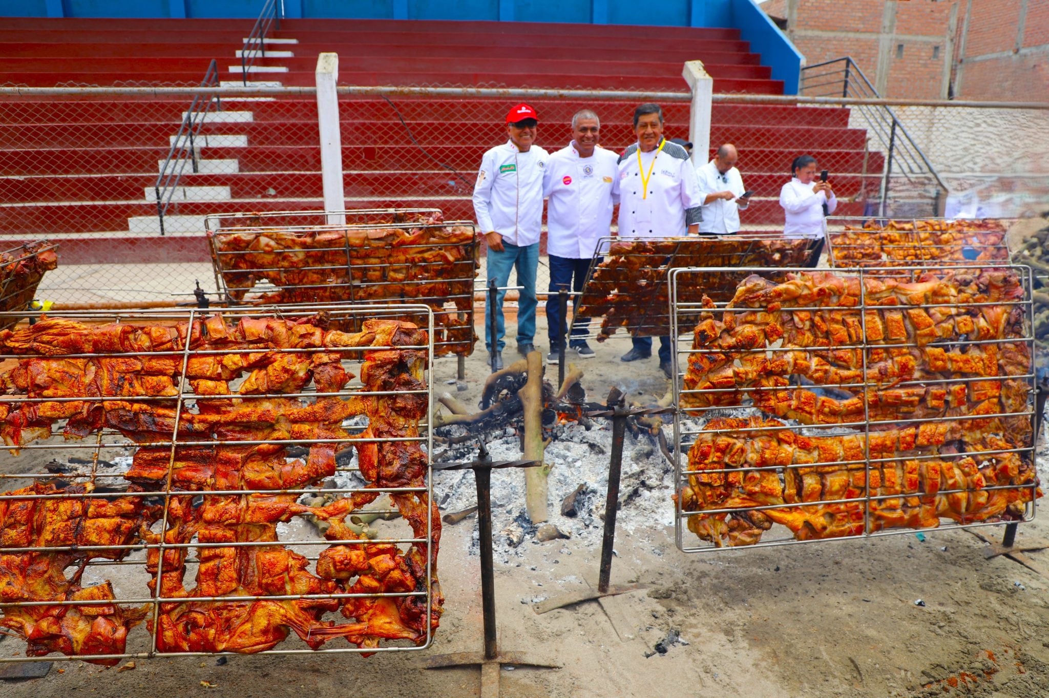 Tres cocineros posan junto a las rejillas de cerdo a las brasas en el Estadio Municipal Rómulo Shaw Cisneros durante la preparación del Festival del Chancho al Palo en Chancay.
