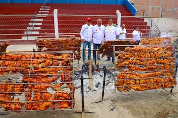 Tres cocineros con uniforme blanco posan detrás de rejillas metálicas con carne de cerdo que se cocina sobre brasas en un estadio