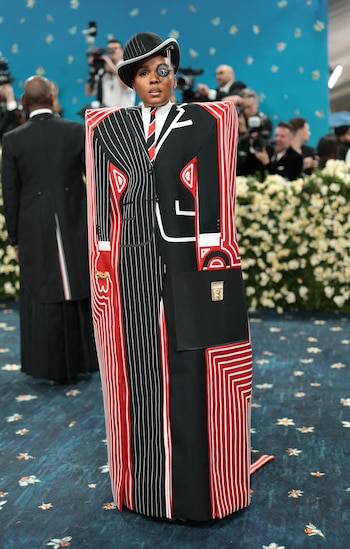 Janelle Monáe llevó un conjunto de silueta escultórica en forma de bloque rectangular, con una superficie que representó la ilustración de un traje masculino a rayas en rojo, negro y blanco. El diseño, altamente estructurado, jugó con la ilusión óptica: integró elementos como solapas, bolsillos y una corbata dibujados o construidos en relieve sobre el propio cuerpo del atuendo. Las rayas verticales y concéntricas reforzaron el efecto gráfico
