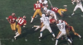 Vista aérea de un partido de fútbol americano con jugadores en uniformes blancos y rojos/dorados. El número 75 de los Miami Dolphins, Manny Fernandez, se prepara para la acción
