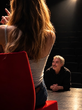 Un hombre con cabello cano mira sonriendo a una mujer de espaldas en una silla roja dentro de un teatro