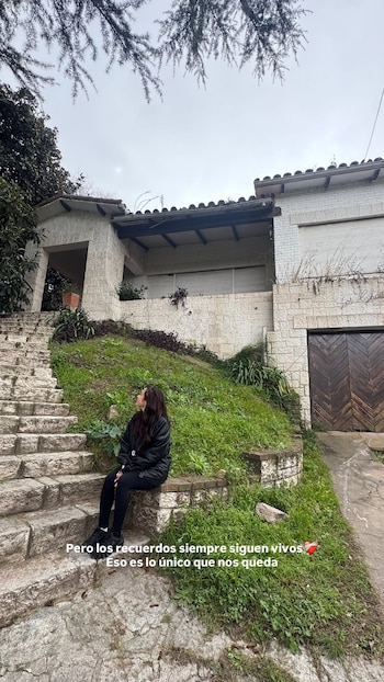 Una mujer sentada en escaleras de piedra con ropa oscura, frente a una casa de fachada de piedra con techo de tejas y un jardín con césped. Cielo nublado