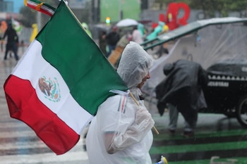 Una persona con impermeable blanco sostiene una bandera de México. Caen gotas de lluvia. Al fondo, personas borrosas y estructuras