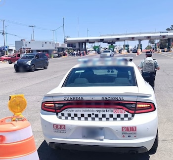 Vista trasera de una patrulla blanca de la Guardia Nacional con un oficial de pie al lado, frente a una caseta de peaje con varios autos. Un cono naranja está en primer plano