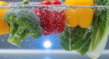 Brócoli, pimientos rojos y amarillos, y bok choy sumergidos en agua con numerosas burbujas, contra un fondo azul claro.