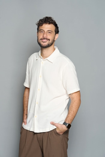 Un hombre de barba y pelo rizado oscuro, con una camisa clara de manga corta y pantalones marrones, sonríe a la cámara con las manos en los bolsillos
