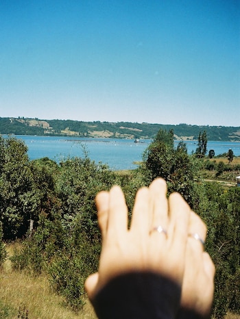 Dos manos entrelazadas en primer plano muestran anillos frente a un lago rodeado de colinas y vegetación