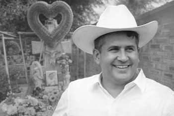 Imagen en blanco y negro de un hombre sonriente con sombrero y camisa clara. Detrás, una tumba o memorial con flores y una corona de corazón.