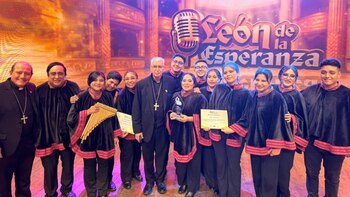 Trece personas, con dos hombres de vestimenta clerical, posan sonrientes en un escenario con logo, sosteniendo diplomas, un trofeo y una zampoña