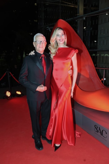 Hombre con traje negro y mujer con vestido rojo satinado posan sonrientes en una alfombra roja junto a una enorme escultura de zapato de tacón rojo