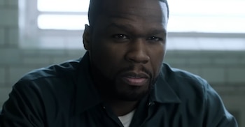 50 Cent con gorra de béisbol negra con el logo de los Yankees, camisa estampada azul y blanca, y pendientes, posa de frente sobre un fondo gris oscuro