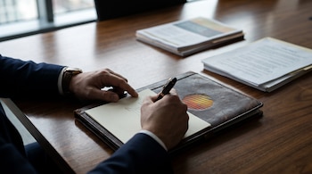 Manos en traje firman un papel con pluma sobre una carpeta de cuero marrón con un emblema circular, sobre una mesa de madera con documentos.