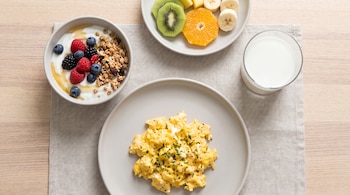 Huevos revueltos en un plato, un cuenco con yogur, frutos rojos y granola, otro plato con kiwi, naranja y plátano, y un vaso de leche.