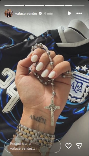 Mano de mujer sostiene rosario plateado con cruz. En la muñeca, un tatuaje que dice 'AMÉN'. Usa pulsera dorada y plateada. Fondo con camiseta de Argentina
