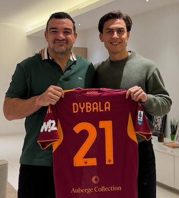 Dos hombres sonríen directamente a la cámara mientras sostienen una camiseta de fútbol color granate con el nombre "DYBALA" y el número "21". El hombre de la izquierda lleva un polo verde y el de la derecha un suéter gris