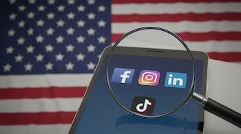 Una lupa amplía los íconos de Facebook, Instagram, LinkedIn y TikTok en la pantalla de un teléfono inteligente, con la bandera de Estados Unidos al fondo.