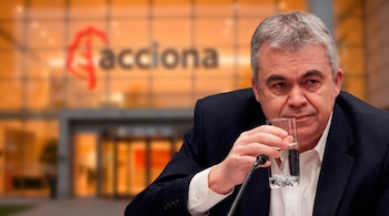 Santos Cerdán, de mediana edad, bebe de un vaso de agua, con un micrófono al lado. El logotipo de Acciona se ve difuminado en el fondo.