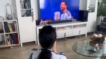 Persona de espaldas con camiseta de Argentina mira un televisor que muestra un jugador de fútbol. Mueble de televisión, estantes con libros y mesa de vidrio en una sala