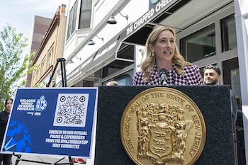 Mujer rubia hablando en un podio con el Gran Sello de Nueva Jersey; a su lado, un cartel azul promociona el programa de recompensas del Mundial