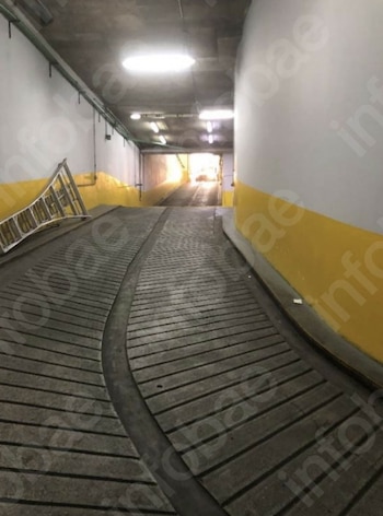 Vista de una rampa de estacionamiento subterráneo de hormigón con paredes blancas y franjas amarillas, una escalera metálica apoyada a la izquierda y luz al fondo