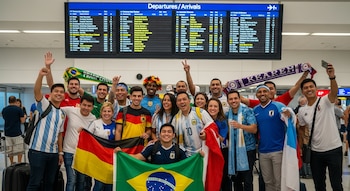 Grupo de entusiastas aficionados al fútbol de diversas nacionalidades posa sonriente con banderas y bufandas en un aeropuerto, con una pizarra de vuelos.
